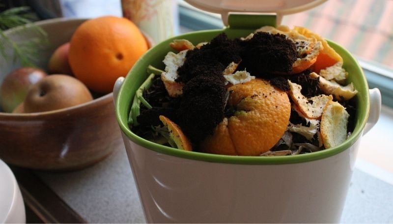 a white plastic kitchen countertop compost bin filled with food scraps including orange peels